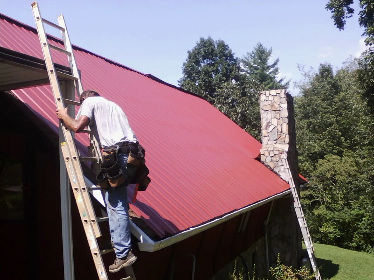 Roofer on a ladder beside a red metal roof and stone chimney — Soffit & Fascia Repair by Roofing Brothers in Greenbelt